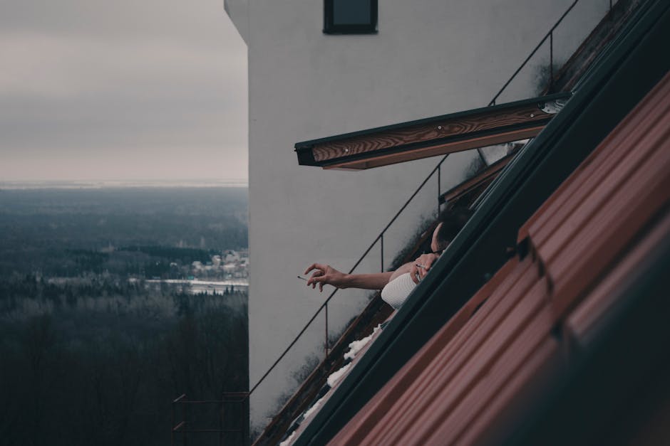 A person enjoys a smoke at a roof window with a view of a sprawling landscape.