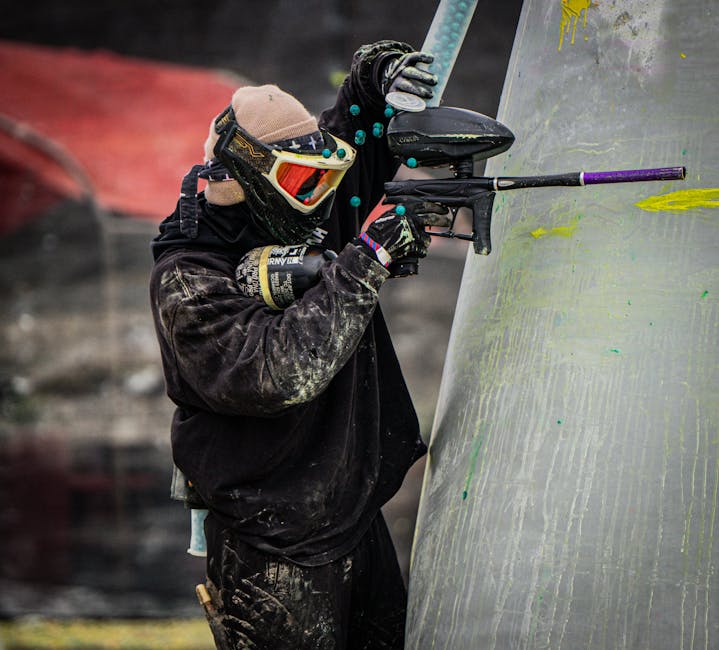 A paintball player in full gear taking aim during a competitive game outdoors.